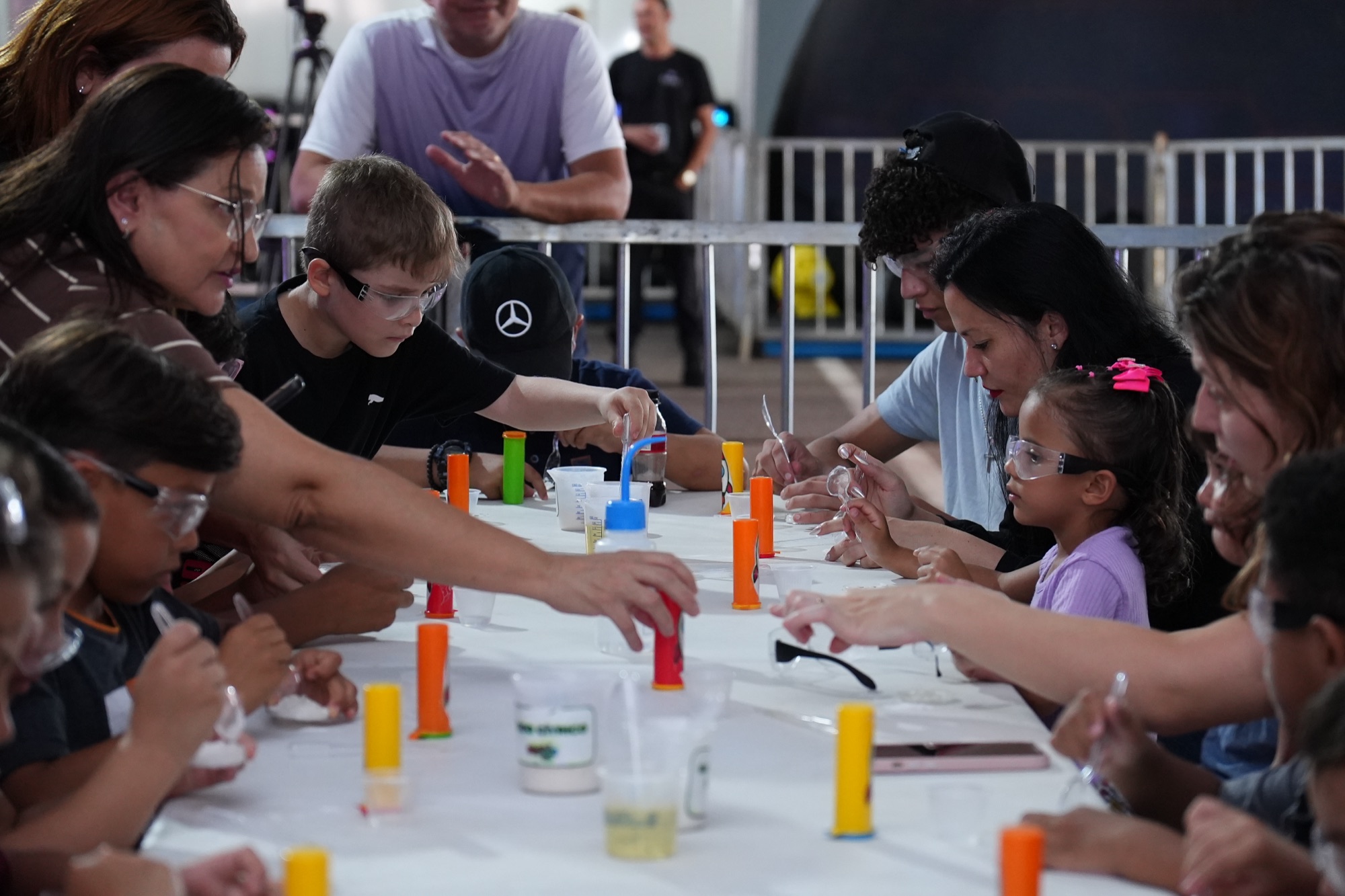 Crianças com óculos de proteção participam de uma experiência científica acompanhadas por adultos