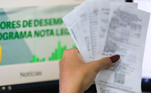Pessoa segura notas fiscais diante de uma tela do programa Nota Legal