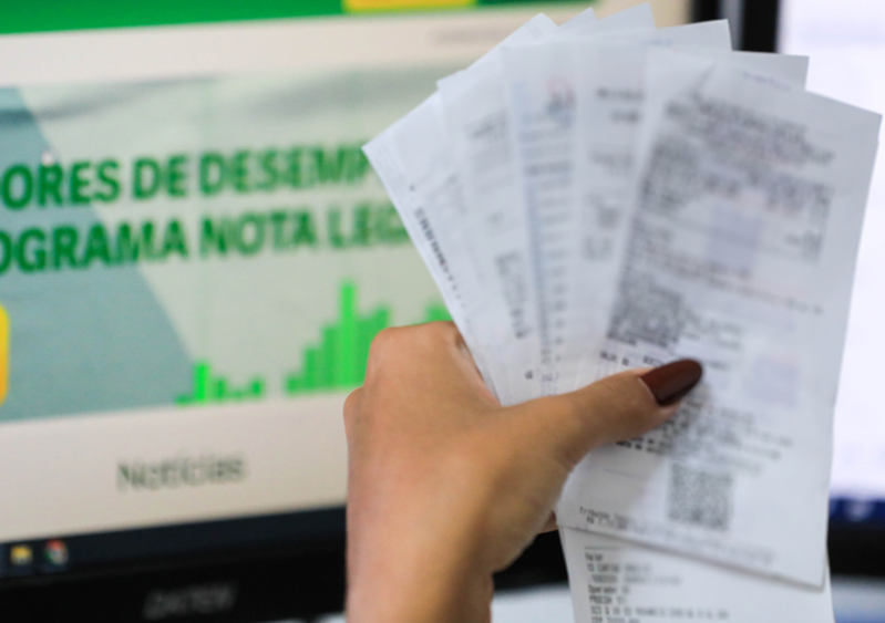 Pessoa segura notas fiscais diante de uma tela do programa Nota Legal