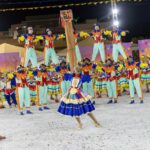 Integrantes de uma quadrilha junina se apresentam com figurinos coloridos em uma arena