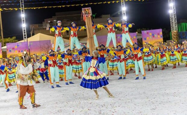 Integrantes de uma quadrilha junina se apresentam com figurinos coloridos em uma arena