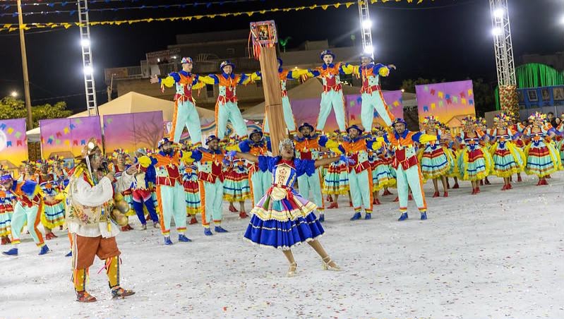 Integrantes de uma quadrilha junina se apresentam com figurinos coloridos em uma arena
