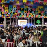 Público participa de festa paroquial com bandeirinhas coloridas, mesas e palco iluminado