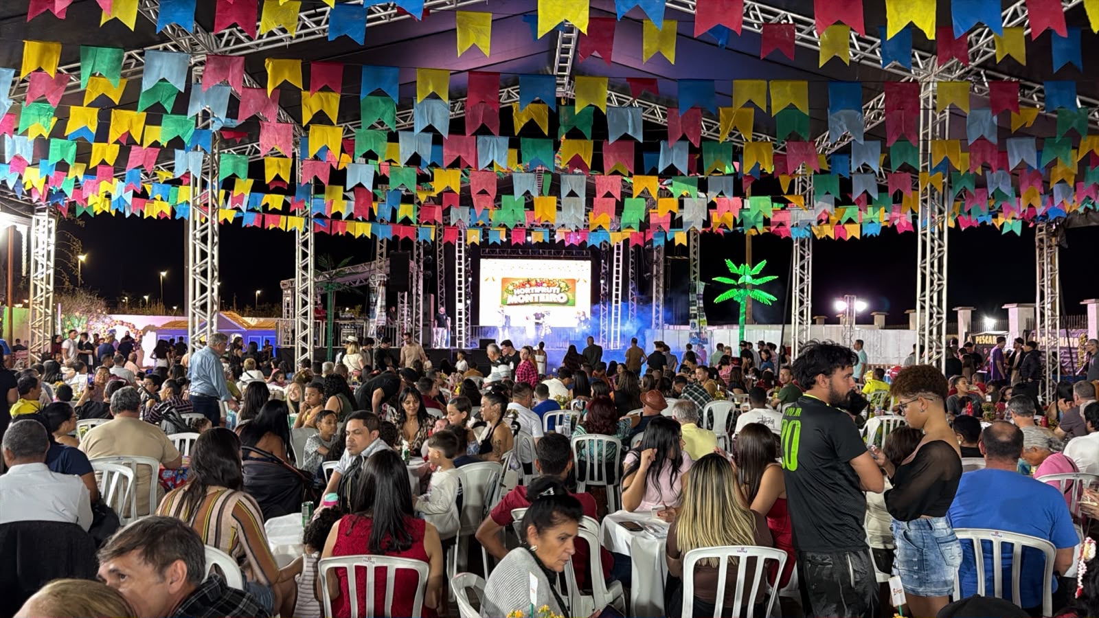 Público participa de festa paroquial com bandeirinhas coloridas, mesas e palco iluminado