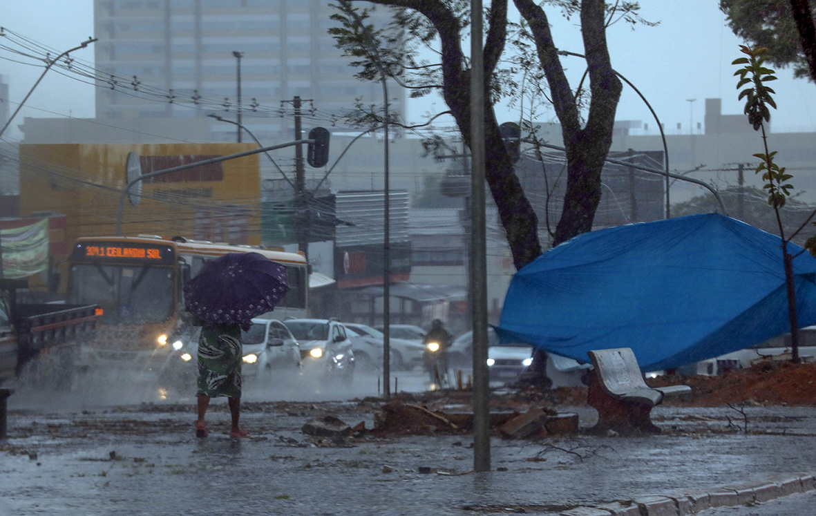 Pedestre com guarda-chuva caminha ao lado de veículos durante chuva intensa no Distrito Federal