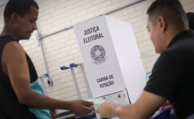 Eleitor entrega documento ao lado de uma cabine de votação
